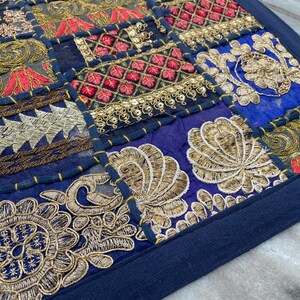 May include: A patchwork textile with a navy blue border. The design features squares of various colors, including navy, red, gold, and gray, with intricate embroidered patterns. The embroidery includes floral and geometric motifs, creating a detailed and textured appearance.