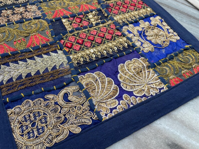 May include: A patchwork textile with a navy blue border. The design features squares of various colors, including navy, red, gold, and gray, with intricate embroidered patterns. The embroidery includes floral and geometric motifs, creating a detailed and textured appearance.