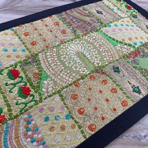 Puede incluir: Textil de patchwork con borde negro. El diseño presenta varios cuadrados bordados en tonos verdes, dorados y crema, con acentos de rojo, azul y naranja. El textil es probablemente un artículo decorativo, como un camino de mesa o un tapiz.