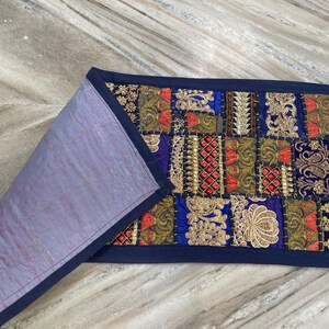 May include: A rectangular, patchwork textile with a navy blue border. The front features a vibrant mix of colors including blue, red, gold, and green, with intricate embroidered patterns. The reverse side is a solid lavender color.
