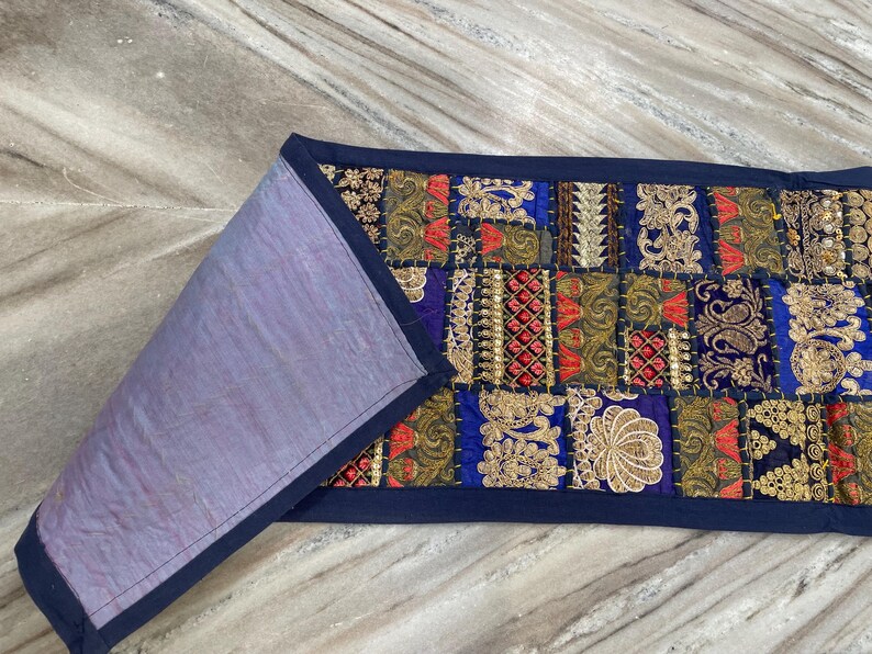 May include: A rectangular, patchwork textile with a navy blue border. The front features a vibrant mix of colors including blue, red, gold, and green, with intricate embroidered patterns. The reverse side is a solid lavender color.