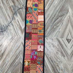 May include: A rectangular patchwork table runner with a black border. The runner features a variety of colorful fabric squares with floral, paisley, and geometric patterns in shades of pink, orange, blue, and gold.