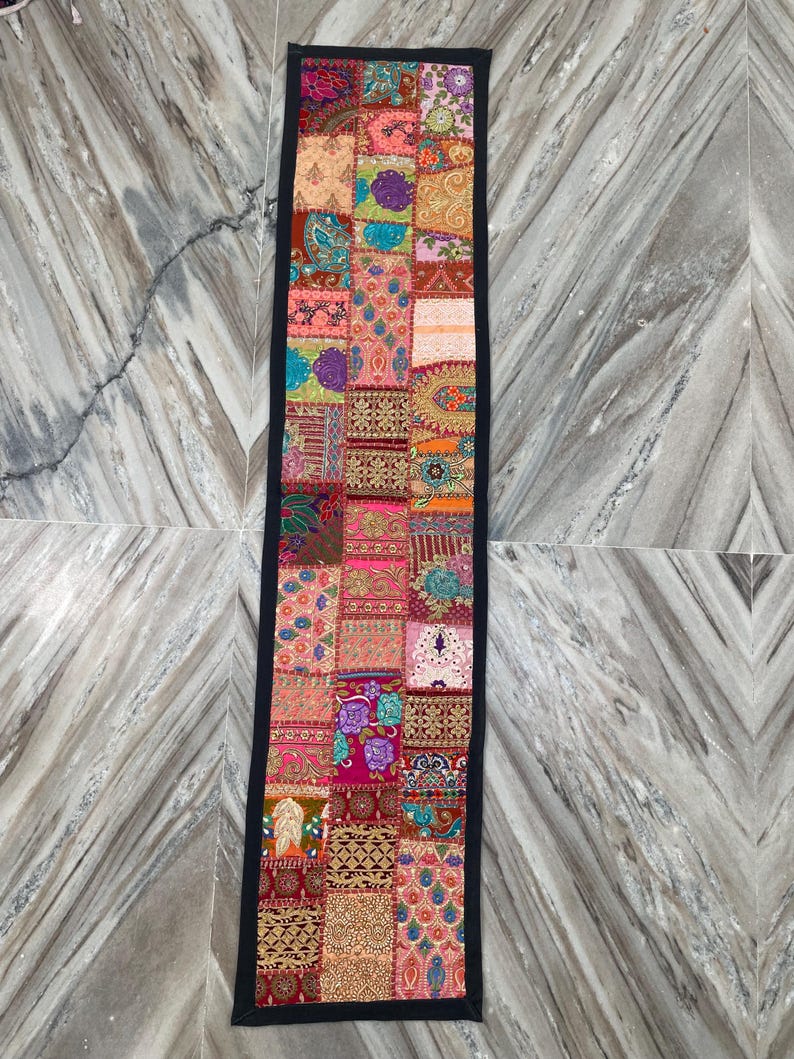 May include: A rectangular patchwork table runner with a black border. The runner features a variety of colorful fabric squares with floral, paisley, and geometric patterns in shades of pink, orange, blue, and gold.