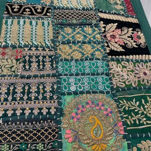May include: A patchwork textile with a variety of embroidered patterns in shades of green, gold, and black. The design includes floral and geometric motifs, with a dark green border. The textile is likely a decorative item, such as a wall hanging or table runner.