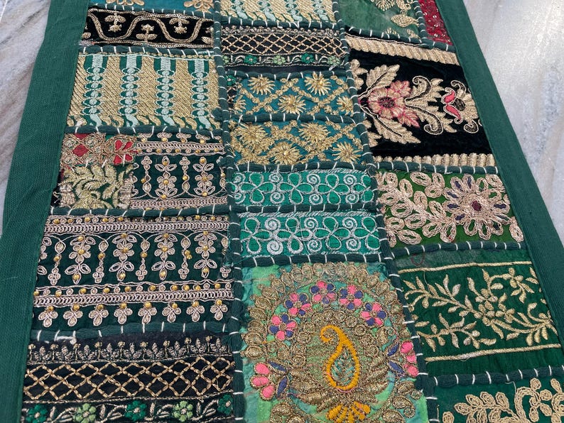 May include: A patchwork textile with a variety of embroidered patterns in shades of green, gold, and black. The design includes floral and geometric motifs, with a dark green border. The textile is likely a decorative item, such as a wall hanging or table runner.