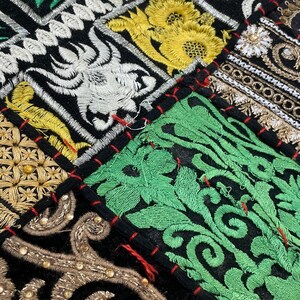 May include: Close-up of a patchwork textile with intricate embroidery. The design features various geometric and floral patterns in gold, green, white, and black. The patches are sewn together with red thread, creating a vibrant and detailed texture.