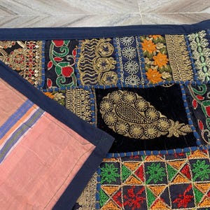 May include: A patchwork textile with a navy blue border. The design incorporates various fabric squares with embroidered patterns in gold, red, orange, and green. A section of pink fabric with blue and orange stripes is visible.