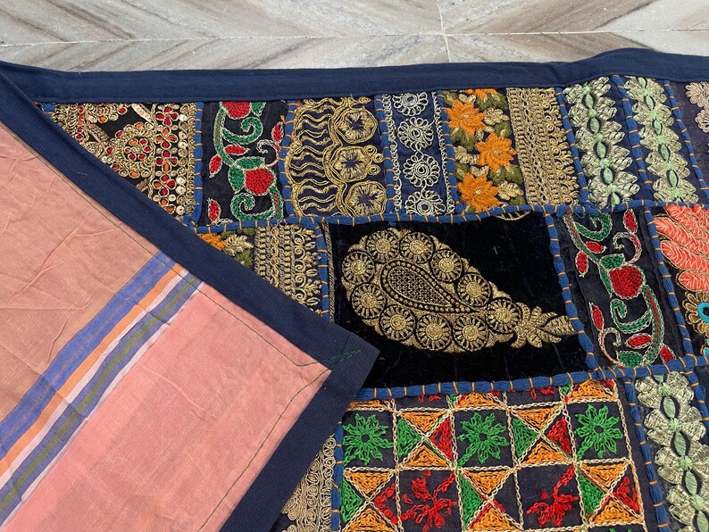 May include: A patchwork textile with a navy blue border. The design incorporates various fabric squares with embroidered patterns in gold, red, orange, and green. A section of pink fabric with blue and orange stripes is visible.