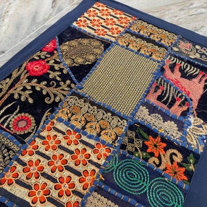 May include: A patchwork textile with a navy blue border. The design features various embroidered squares with floral and geometric patterns in shades of orange, gold, red, and green on a black background. The textile is likely a decorative item.