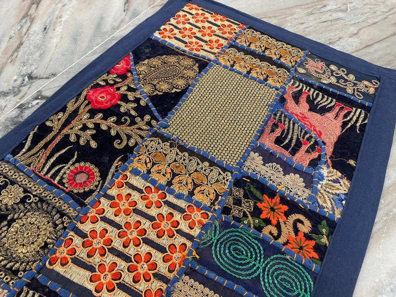 May include: A patchwork textile with a navy blue border. The design features various embroidered squares with floral and geometric patterns in shades of orange, gold, red, and green on a black background. The textile is likely a decorative item.