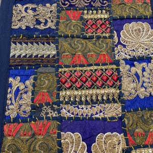 May include: A patchwork textile with a navy blue border. The design features embroidered squares in shades of blue, gold, red, and green. Intricate floral and geometric patterns adorn each square, creating a rich, textured appearance.