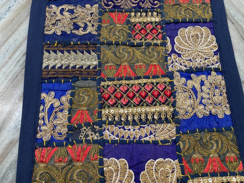 May include: A patchwork textile with a navy blue border. The design features embroidered squares in shades of blue, gold, red, and green. Intricate floral and geometric patterns adorn each square, creating a rich, textured appearance.