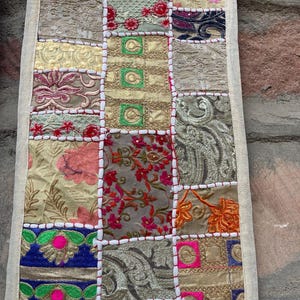 May include: A rectangular patchwork textile with a variety of patterned squares in shades of gold, pink, blue, and green. The squares are stitched together with white thread, and the edges are finished with a cream-colored border. The textile has an overall vintage aesthetic.