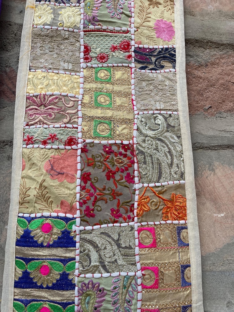 May include: A rectangular patchwork textile with a variety of patterned squares in shades of gold, pink, blue, and green. The squares are stitched together with white thread, and the edges are finished with a cream-colored border. The textile has an overall vintage aesthetic.