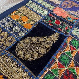 May include: Close-up of a patchwork textile with intricate embroidery. The design features a variety of patterns, including floral and paisley motifs, in shades of gold, orange, green, red, and blue. The textile is likely a decorative item, such as a throw or wall hanging.
