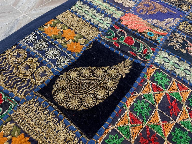 May include: Close-up of a patchwork textile with intricate embroidery. The design features a variety of patterns, including floral and paisley motifs, in shades of gold, orange, green, red, and blue. The textile is likely a decorative item, such as a throw or wall hanging.