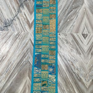 May include: A rectangular, turquoise-bordered table runner. The runner features a patchwork design with various patterns in gold, teal, and blue. The design includes floral and geometric motifs, creating a decorative textile.