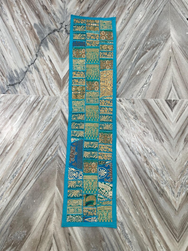 May include: A rectangular, turquoise-bordered table runner. The runner features a patchwork design with various patterns in gold, teal, and blue. The design includes floral and geometric motifs, creating a decorative textile.