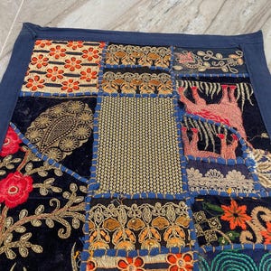 May include: A patchwork textile with a navy blue border. The design features various squares with floral, paisley, and geometric patterns in shades of red, orange, gold, and blue. The textile is likely made of velvet and embroidered with gold thread.