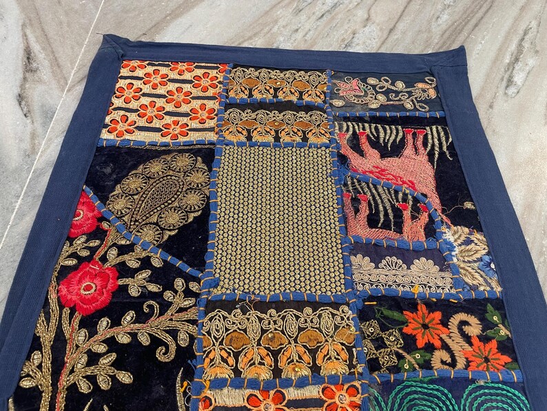 May include: A patchwork textile with a navy blue border. The design features various squares with floral, paisley, and geometric patterns in shades of red, orange, gold, and blue. The textile is likely made of velvet and embroidered with gold thread.