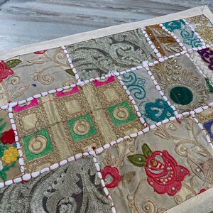 May include: A rectangular patchwork textile with a cream-colored border. The design features various embroidered squares in shades of gold, green, pink, and blue, with floral and geometric patterns. The textile is likely used as a decorative element.