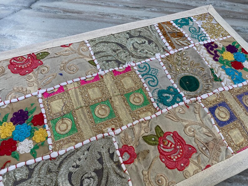 May include: A rectangular patchwork textile with a cream-colored border. The design features various embroidered squares in shades of gold, green, pink, and blue, with floral and geometric patterns. The textile is likely used as a decorative element.