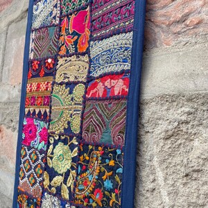 May include: A rectangular wall hanging with a patchwork design. The piece features a dark blue border and a variety of embroidered fabric squares in vibrant colors like pink, orange, and gold. The design includes floral and geometric patterns.