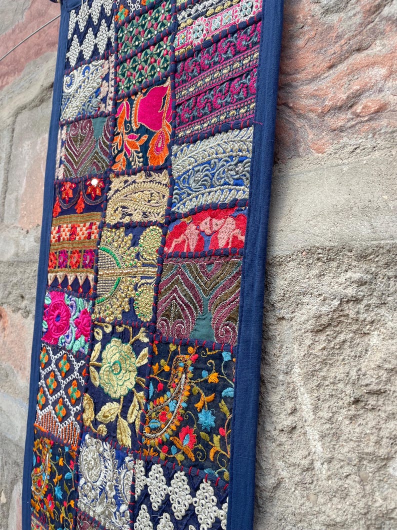 May include: A rectangular wall hanging with a patchwork design. The piece features a dark blue border and a variety of embroidered fabric squares in vibrant colors like pink, orange, and gold. The design includes floral and geometric patterns.
