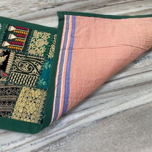 May include: A decorative textile with a patchwork design. The front features embroidered patches in green, black, and gold, with intricate patterns. The reverse side is a solid peach color. The edges are trimmed with a dark green border.