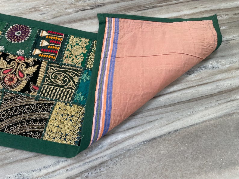 May include: A decorative textile with a patchwork design. The front features embroidered patches in green, black, and gold, with intricate patterns. The reverse side is a solid peach color. The edges are trimmed with a dark green border.