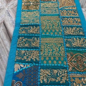 May include: A rectangular, patchwork textile in shades of teal, gold, and blue. The design features embroidered floral and geometric patterns. A bright teal border frames the edges of the textile, which appears to be a table runner.