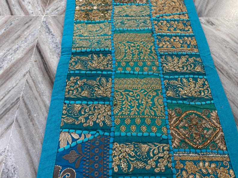 May include: A rectangular, patchwork textile in shades of teal, gold, and blue. The design features embroidered floral and geometric patterns. A bright teal border frames the edges of the textile, which appears to be a table runner.