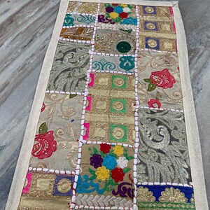 May include: A rectangular patchwork textile with a cream-colored border. The patchwork features squares of various colors and patterns, including floral embroidery, gold fabric, and embroidered details. The textile is likely a decorative item.