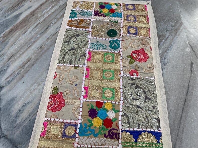 May include: A rectangular patchwork textile with a cream-colored border. The patchwork features squares of various colors and patterns, including floral embroidery, gold fabric, and embroidered details. The textile is likely a decorative item.