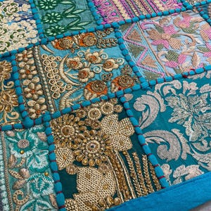 May include: Close-up of a patchwork textile with intricate embroidery. The design features squares of various colors, including teal, gold, pink, and cream. Detailed floral and geometric patterns are visible, with a teal border.