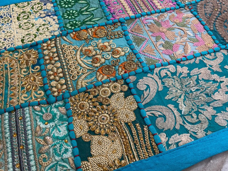 May include: Close-up of a patchwork textile with intricate embroidery. The design features squares of various colors, including teal, gold, pink, and cream. Detailed floral and geometric patterns are visible, with a teal border.