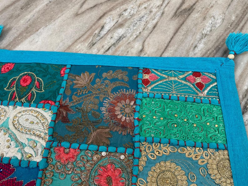 May include: A patchwork textile with a vibrant mix of colors and patterns. The design features squares of teal, turquoise, and gold fabric with floral and paisley embroidery. A bright blue border and tassels complete the look.