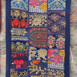 May include: A rectangular textile with a patchwork design. The patches feature intricate embroidery in shades of blue, pink, orange, and gold. The edges are trimmed with a dark blue border. The textile is held by a black clip.