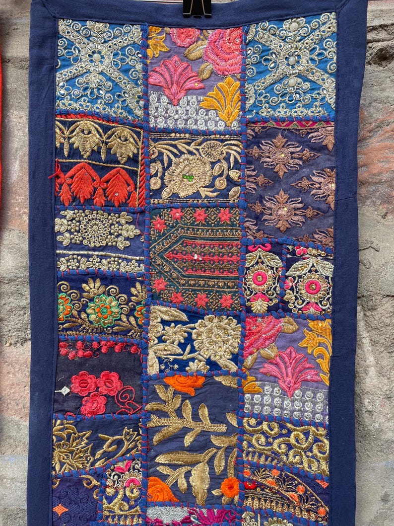 May include: A rectangular textile with a patchwork design. The patches feature intricate embroidery in shades of blue, pink, orange, and gold. The edges are trimmed with a dark blue border. The textile is held by a black clip.