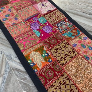 May include: A rectangular patchwork table runner with a black border. The runner features a variety of colorful fabric squares with intricate embroidery and embellishments in shades of pink, red, gold, and teal. The design is a mix of floral and geometric patterns.