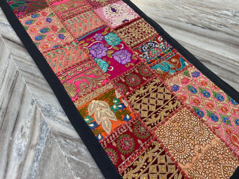 May include: A rectangular patchwork table runner with a black border. The runner features a variety of colorful fabric squares with intricate embroidery and embellishments in shades of pink, red, gold, and teal. The design is a mix of floral and geometric patterns.