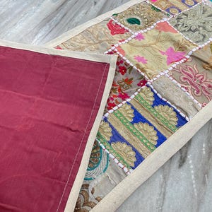 May include: A patchwork table runner with a burgundy backing. The top features a variety of fabric squares in floral and geometric patterns, including pink, blue, and gold. The edges are trimmed with a cream-colored border.