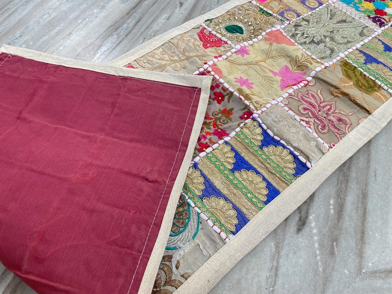 May include: A patchwork table runner with a burgundy backing. The top features a variety of fabric squares in floral and geometric patterns, including pink, blue, and gold. The edges are trimmed with a cream-colored border.