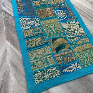May include: A rectangular patchwork textile with a turquoise border. The patchwork design features squares of teal, gold, and blue fabrics with embroidered floral and geometric patterns. The textile is likely a table runner or decorative accent.