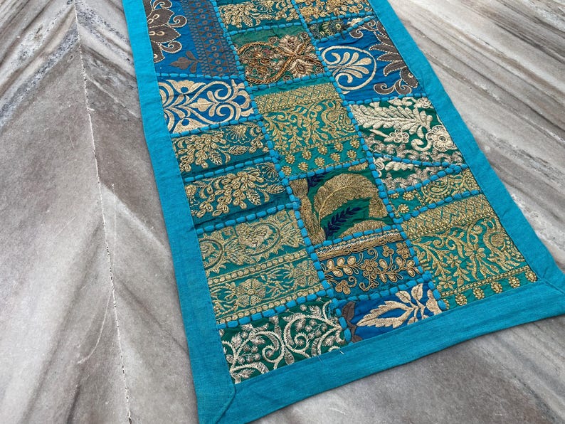 May include: A rectangular patchwork textile with a turquoise border. The patchwork design features squares of teal, gold, and blue fabrics with embroidered floral and geometric patterns. The textile is likely a table runner or decorative accent.