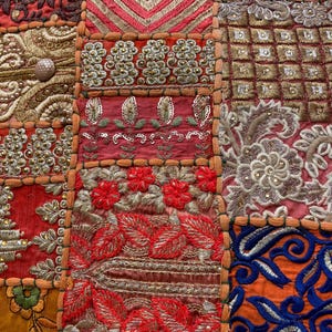 May include: Close-up of a patchwork textile with intricate embroidery. The design features a variety of colors including red, orange, gold, and blue. The textile is divided into squares with floral and geometric patterns.