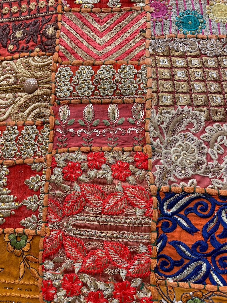 May include: Close-up of a patchwork textile with intricate embroidery. The design features a variety of colors including red, orange, gold, and blue. The textile is divided into squares with floral and geometric patterns.