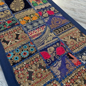 May include: A patchwork textile with a navy blue border. The design features squares with intricate embroidery in gold, red, orange, and blue. The textile has a vintage, bohemian aesthetic.
