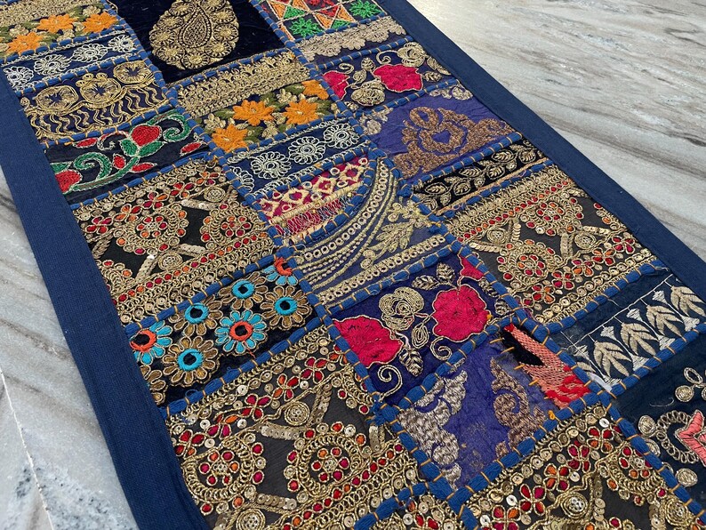 May include: A patchwork textile with a navy blue border. The design features squares with intricate embroidery in gold, red, orange, and blue. The textile has a vintage, bohemian aesthetic.