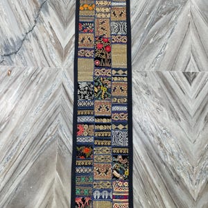 May include: A long, rectangular patchwork textile with a black border. The patchwork design features a variety of colorful, embroidered squares with floral and geometric patterns. The textile is likely a table runner or wall hanging.